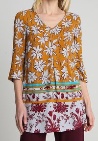 Tunique moutarde avec motif floral blanc, col en V, manches trois-quarts, accents rayés rouge et bleu, et un design floral contrastant en bas.
