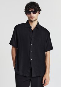 Mann mit dunklem lockigem Haar, der schwarze Sonnenbrille trägt und ein lockeres schwarzes kurzärmliges Hemd mit Knöpfen und schwarze Hosen anhat, steht vor einem weißen Hintergrund.