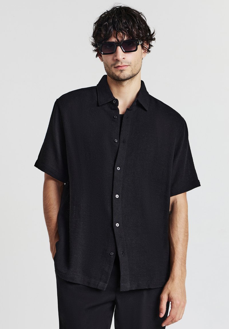 Mann mit dunklem lockigem Haar, der schwarze Sonnenbrille trägt und ein lockeres schwarzes kurzärmliges Hemd mit Knöpfen und schwarze Hosen anhat, steht vor einem weißen Hintergrund.