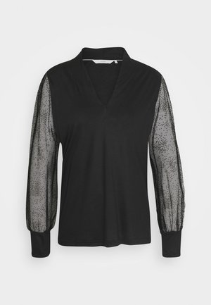 Blouse noire à manches longues avec manches en maille transparente à pois, col en V profond et poignets unis sur fond blanc uni.