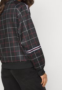 Černá mikina se mřížkovaným vzorem v bílé a červené barvě, žebrované manžety a pruhovaný akcent na rukávu. Hladká, lehká tkanina.