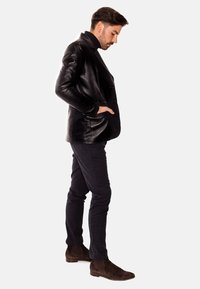 Chaqueta de cuero negro de corte cruzado con una textura suave, bolsillos laterales, llevada sobre pantalones oscuros ajustados y botas de ante marrón hasta el tobillo.