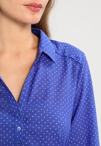 Camicia blu a bottoni realizzata in tessuto leggero, caratterizzata da un motivo di piccoli triangoli bianchi, con un colletto strutturato e una texture liscia.