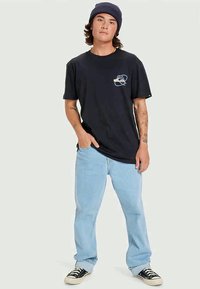 Juodas medvilninis marškinėliai su mažą grafika ant krūtinės, derinami su šviesiai mėlynomis džinsinėmis kelnėmis ir juodais sportbačiais. Lygus audinys, laisvas modelis.