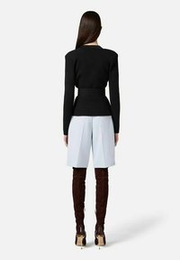 Donna con capelli lunghi e scuri che indossa una maglietta nera aderente a maniche lunghe, pantaloncini grigio chiaro fino al ginocchio e stivali marrone scuro sopra il ginocchio con tacco.