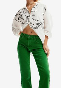 Camisa blanca de botonadura con texto manuscrito en negro, combinada con unos jeans verdes de cintura alta. La camisa tiene las mangas arremangadas; los jeans son lisos y ajustados.