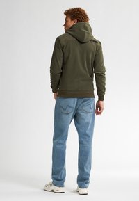 Olijfgroen hoodie met een gestructureerde afwerking, voorzien van een capuchon en geribbelde manchetten, gecombineerd met lichtblauwe jeans en witte sneakers.