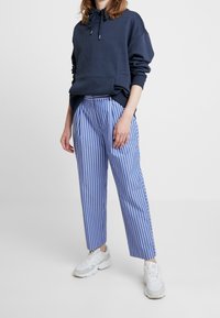 Granatowy hoodie z przednią kieszenią, zestawiony z niebieskimi i białymi spodniami w paski o tapered cut oraz białymi trampkami, oferujący luźny krój i sznurek do regulacji.