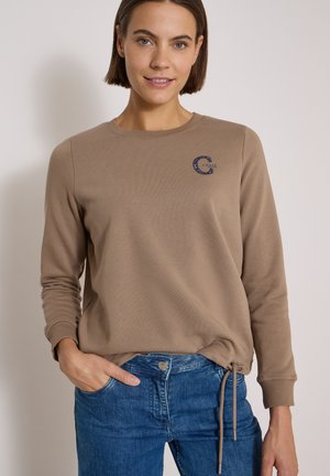 Kvinde med kort brunt hår iført en beige langærmet sweatshirt med et lille logo og blå jeans, står med den ene hånd i lommen.