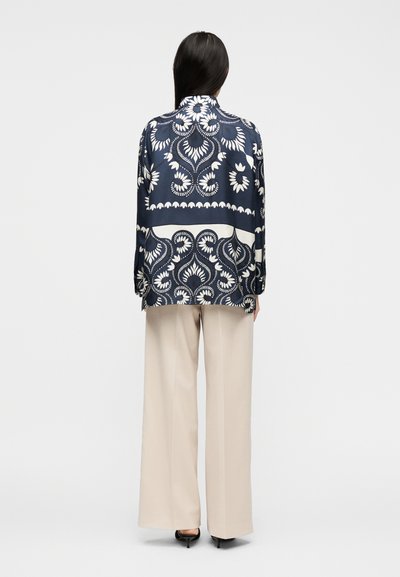 Γυναίκα με μακριά σκούρα μαλλιά, φοράει μπλούζα με σχέδια σε ναυτικό μπλε και άσπρο και μπεζ παντελόνι με φαρδιά πόδια, στέκεται γυρισμένη προς τα πίσω σε απλό φόντο.