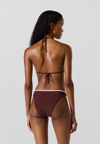Hnedý bikini set s halterneck a zaväzovaním na chrbte. Spodný diel má zúbkovaný biely lem pozdĺž pásu. Hladký, pružný materiál.