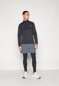 Top de sport à manches longues et fermeture éclair noir en tissu texturé ; shorts gris ; leggings noirs ; chaussures de course blanches avec des accents. Logo New Balance visible.