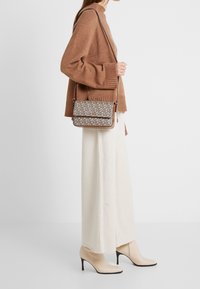 Hnědá crossbody taška s vzorovaným předním dílem, hladkou texturou a hnědým koženým lemem. Spárována s béžovým svetrem a širokými krémovými kalhotami.