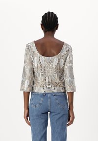 Spitzenoberteil mit einem beigen und silbernen Blumenmuster, Dreiviertelärmel und einer runden Rückenpartie, kombiniert mit blauen High-Waist-Jeans.