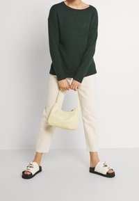 Waldgrüner Pullover, cremefarbene, schmal geschnittene Hose, weiße Rutschen-Sandalen mit Schleife und eine kleine gelbe Tasche. Strukturierter Strick und glattes Taschenmaterial.