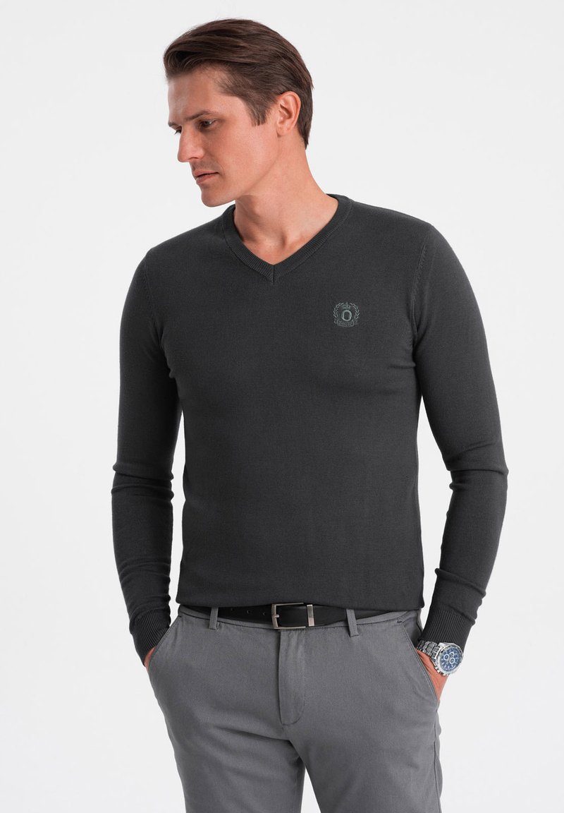 Dunkelgrauer, langärmeliger V-Ausschnitt-Pullover mit gerippten Bündchen und einem kleinen Logo-Detail auf der Brust, kombiniert mit hellgrauen Hosen und einer Uhr.