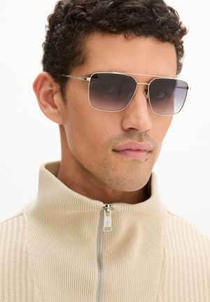 Man draagt een beige ribgebreide sweater met ritssluiting en grote vierkante donkere zonnebril, kijkt iets zijwaarts.