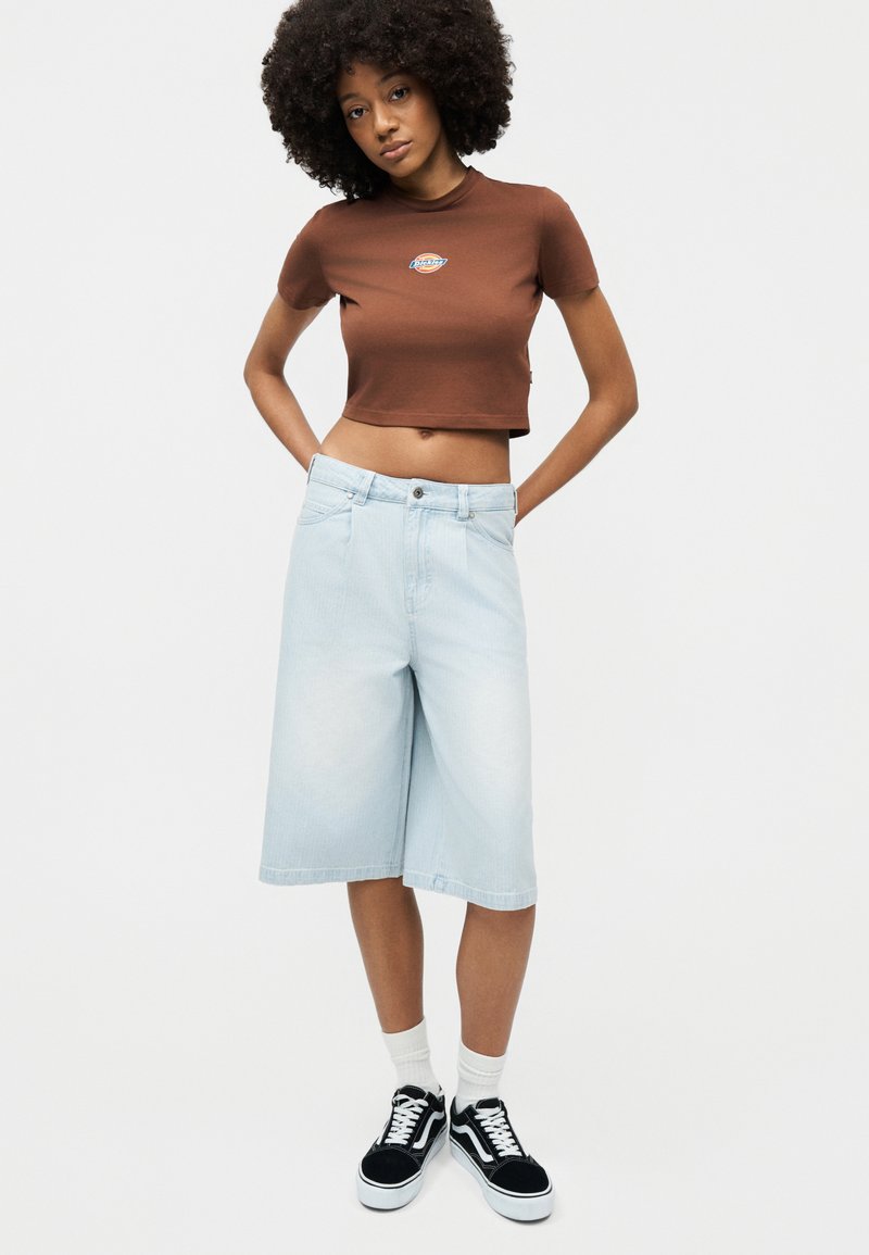 Donna con capelli ricci che indossa una maglietta marrone corta, pantaloncini di jeans azzurri fino al ginocchio, calzini bianchi e scarpe da ginnastica nere, in piedi con le mani dietro la schiena.