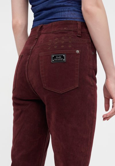 Personne portant un pantalon rouge foncé avec une étiquette noire sur la poche arrière et un haut bleu, montré de la taille au milieu de la cuisse.