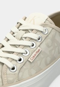Primo piano di una sneaker beige Calvin Klein con lacci bianchi, occhielli argentati, etichetta in tessuto con brand e grande logo "cK" sulla linguetta.