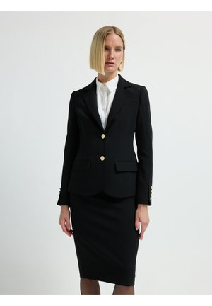 Blazer nero con due tasche frontali e bottoni dorati, abbinato a una gonna nera aderente. Il tessuto sembra liscio e strutturato.