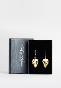 Guldbladformade örhängen visas i en svart låda med texturerad yta och logotyp, med intrikata utskurna detaljer.