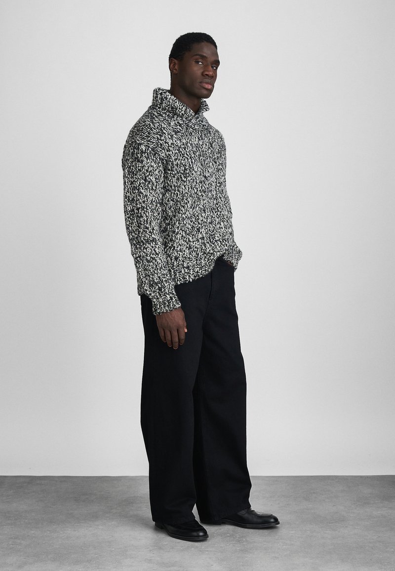 Maglione spesso in bianco e nero a maglia con colletto alto, abbinato a pantaloni neri larghi e scarpe nere, su uno sfondo neutro.