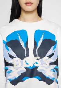 Personne portant une chemise blanche avec un grand motif floral abstrait bleu, noir et gris clair, avec "BOSS" brodé en blanc sur la poitrine.