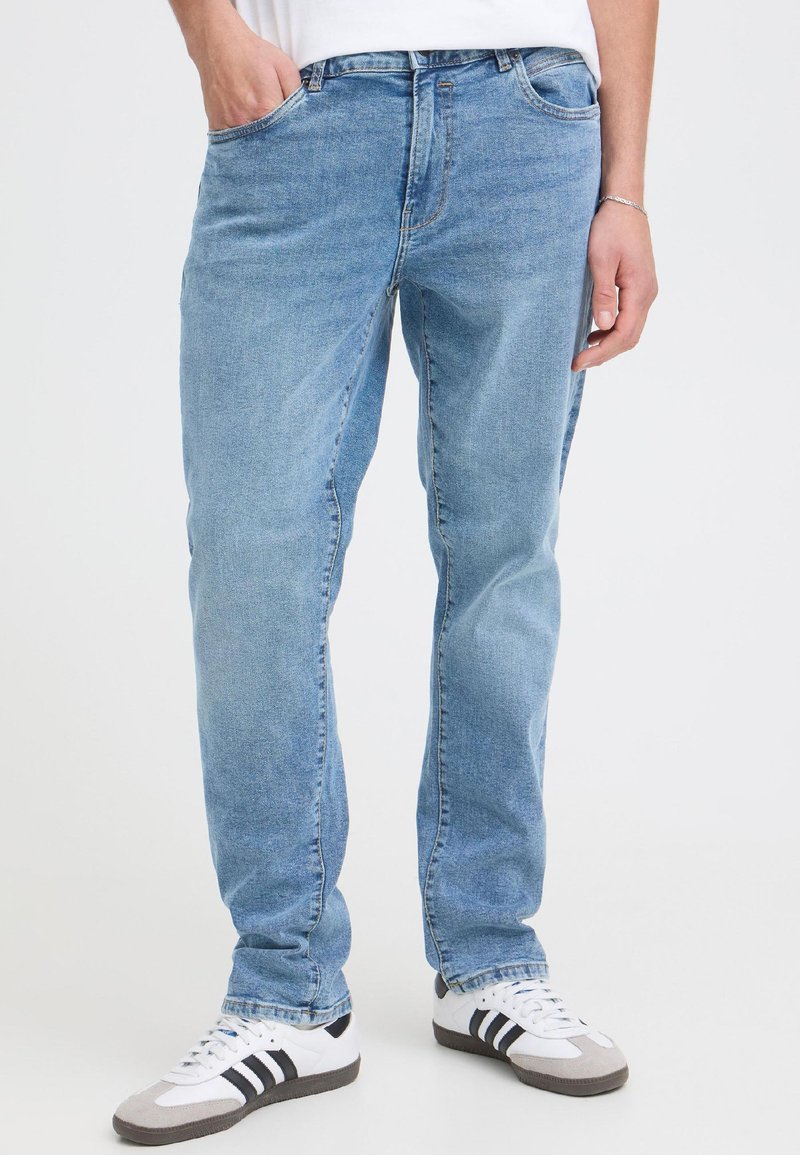 Mann trägt hellblaue Jeans mit geradem Bein und weiße Sneaker mit schwarzen Streifen, steht mit einer Hand in der Tasche.