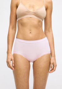 Vrouw in een huidskleurige beha en lichtroze high-waist onderbroek met kant, staande tegen een eenvoudige witte achtergrond.