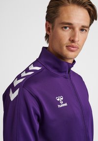 Giacca zip-up viola con colletto alto, caratterizzata da motivi a chevron bianchi sulle spalle e un logo sul petto. Texture liscia.