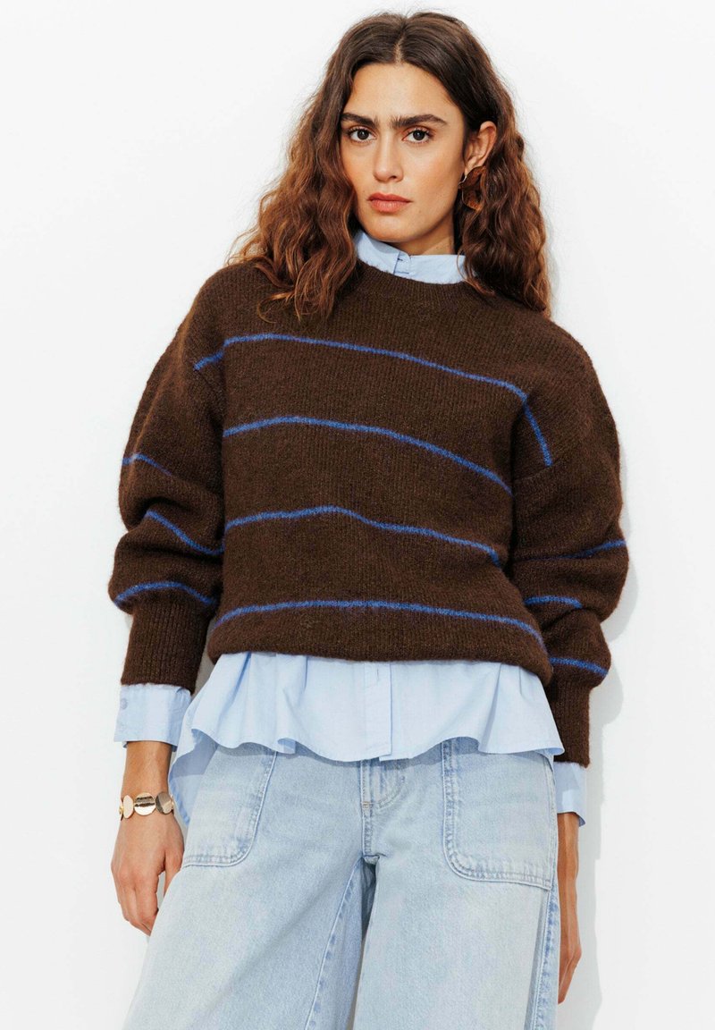 Pull en maille marron floue avec des rayures bleues, associé à une chemise bleu clair à col, et un jean en denim clair à jambes larges. Les accessoires incluent un bracelet.