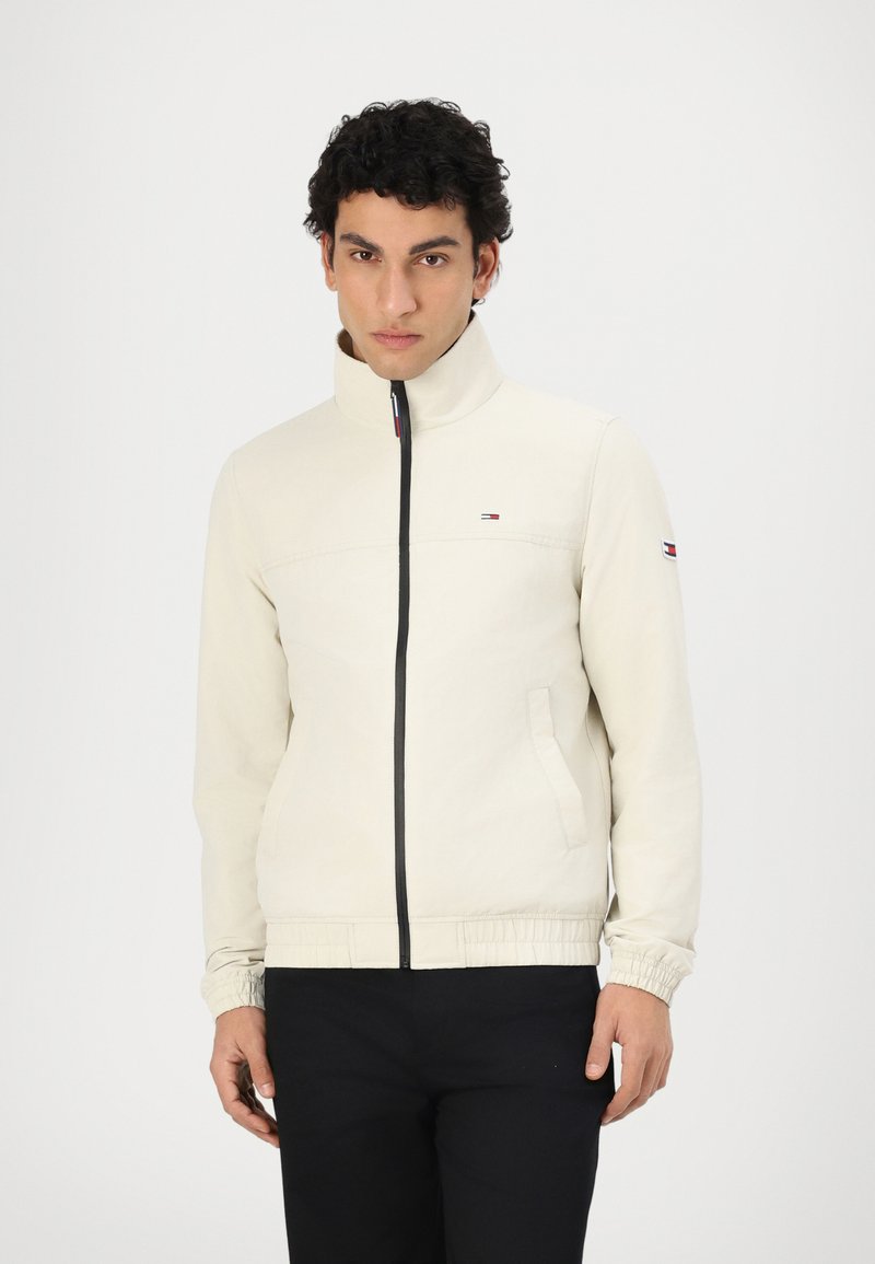 Cream-colored jacket with a high collar, black zipper, and elastic cuffs. Features a small logo on the chest and side pockets.