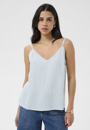 Femme aux cheveux foncés portant des boucles d'oreilles créoles dorées, un haut blanc texturé sans manches, un jean bleu et un bracelet doré au poignet gauche.