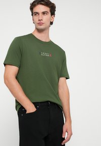 Πράσινο βαμβακερό t-shirt με το λογότυπο "TOMMY HILFIGER" σε λευκό και κόκκινο, στρογγυλή λαιμόκοψη και κοντά μανίκια, σε συνδυασμό με μαύρα τζιν.