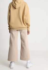 Sudadera con capucha beige de corte relajado, combinada con pantalones anchos de un beige claro y zapatillas blancas, vista desde la parte posterior.