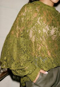 Oliven grøn blonde top med blomster mønstre, gennemsigtig tekstur og brede ærmer. Beklædningen ser ud til at være lagdelt over en tætsiddende grå top.
