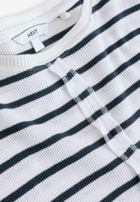 Tissu gaufré blanc avec des rayures horizontales noires et une patte de boutonnage à l'avant, avec une étiquette de marque "NEXT" au col.