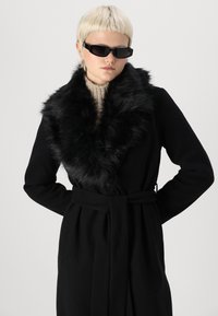 Sort frakke med falsk pelskrave, bælte i taljen og lange ærmer. Båret over en beige strikket sweater med en struktureret stof.
