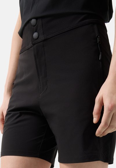 Nahaufnahme einer Person, die schwarze, hoch taillierte Shorts mit zwei vorderen Knöpfen und einer seitlichen Reißverschlusstasche trägt und mit entspannten Händen steht.