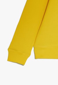 Manga y dobladillo de sudadera amarilla con puños acanalados y detalles de costura sobre un fondo blanco.