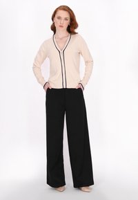 Cremefarbener Cardigan mit marineblauen Akzenten und Knopfverschluss. Kombiniert mit schwarzen, weit geschnittenen Hosen und nudefarbenen High Heels, was eine schlanke, moderne Silhouette zeigt.