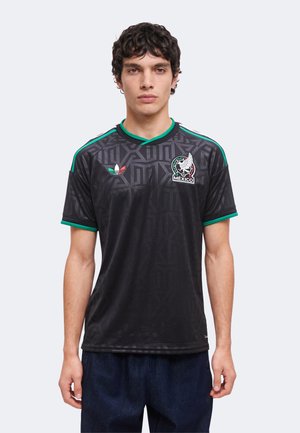 Mladi muškarac nosi crnu nogometnu majicu Meksika s zelenim obrubom, Adidasovim logotipom i grbom momčadi Meksika, stojeći na jednobojnoj pozadini.