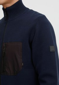 Sudadera navy con cremallera y cuello acanalado, que presenta un bolsillo en el pecho en tejido oscuro y un pequeño parche con logo negro en la manga.