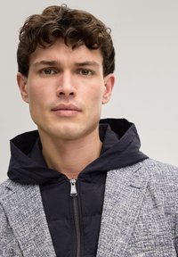 Jonge man met krullend bruin haar, gekleed in een zwarte jas met rits en capuchon onder een grijs geruit colbert, kijkt recht vooruit met een neutrale uitdrukking.