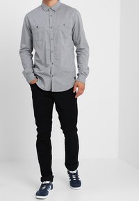 Chemise grise à boutons avec manches longues, deux poches sur la poitrine, portée avec un pantalon noir et des baskets bleu marine. Matière en coton, design uni.