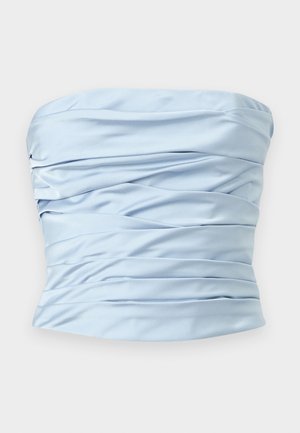 Femme portant un haut bleu clair froncé sans bretelles, une longue jupe blanche et des talons noirs à lacets, debout avec une main sur la hanche.