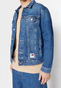 Sininen farkkutakki metallinapeilla, kaksi rintataskua ja alareunassa logoaiheinen kangasmerkki. Siinä on klassinen kaulus ja erottuvat kontrastitikkaukset.