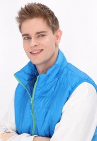 Gilet imbottito blu chiaro con colletto alto e dettagli verdi della cerniera, indossato sopra una maglietta bianca a maniche lunghe. Tessuto a trapunta a rombi incrociati.