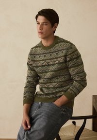 Fiatal férfi mintás zöld és bézs pulóverben és szürke farmerban ül egy széken, egyik kezét a zsebébe dugva, egy sima háttér előtt.